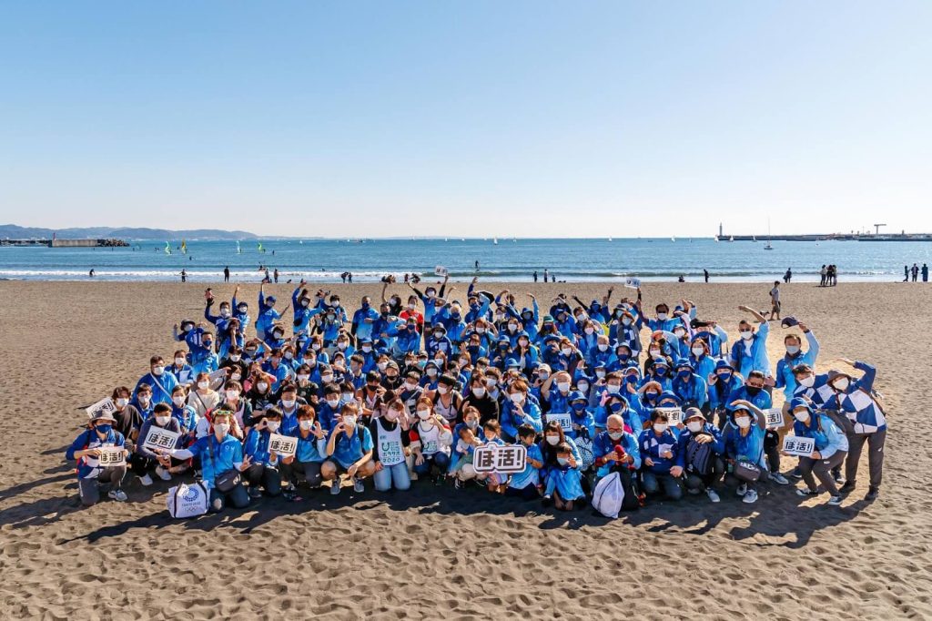 青いユニフォームを着て海岸で集合写真を撮るゴミ拾いボランティアの様子