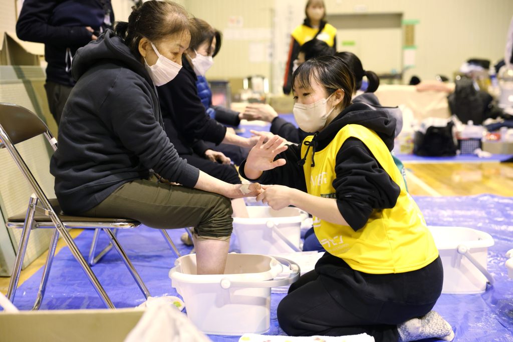 地震の避難所で足湯を利用する人の手をさすりながら会話するボランティアたち