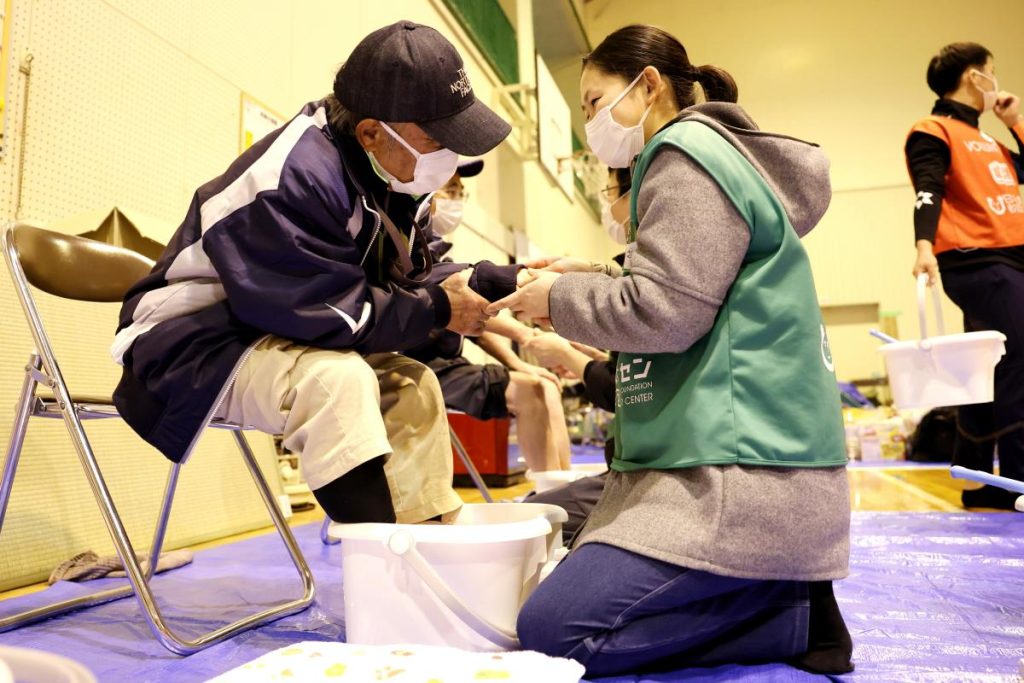 地震の避難所で足湯を利用する人の手をさすり、目を合わせて会話するボランティア