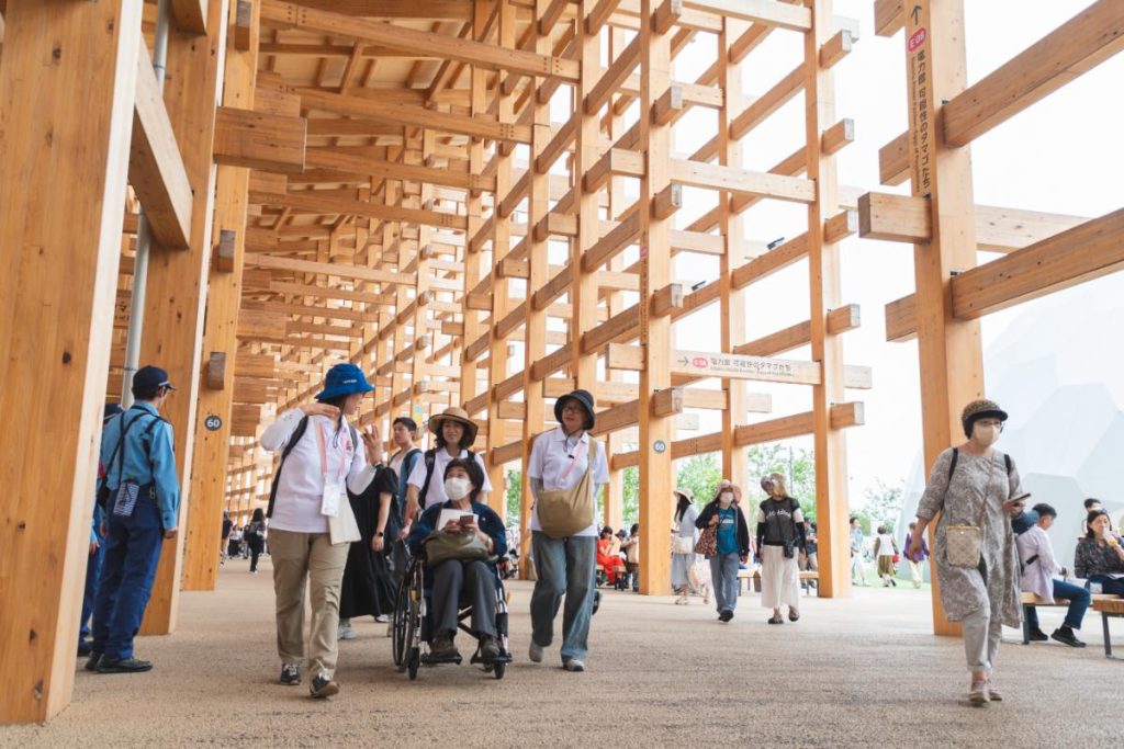 大阪・関西万博会場内の大屋根リングの下で案内をするLET’S EXPOの会場内サポートボランティアと車いすユーザー