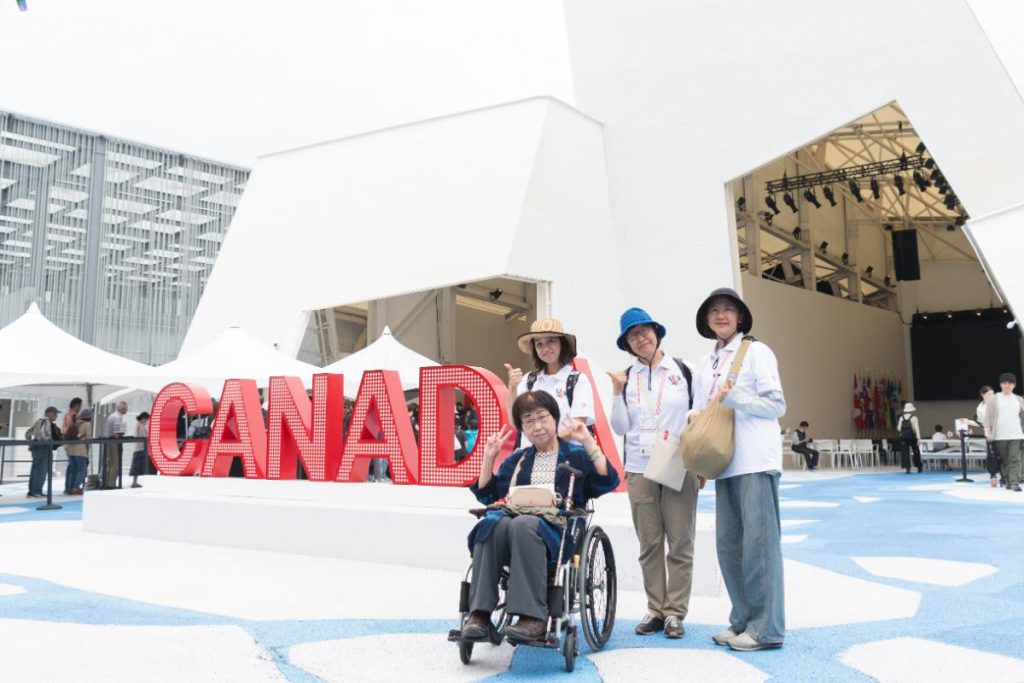 カナダ館パビリオン前で集合写真を撮る車いすユーザーとLET’S EXPOの会場内サポートボランティア